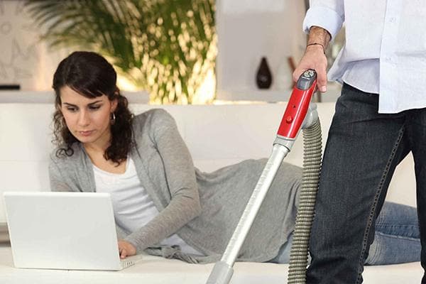 Femme allongée sur un canapé avec un ordinateur portable pendant que l'homme passe l'aspirateur
